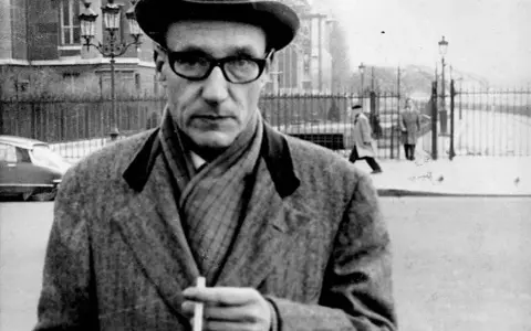 William Burroughs, con cappello e occhiali, tiene una sigaretta in mano, passeggiando in una strada urbana con persone sullo sfondo.