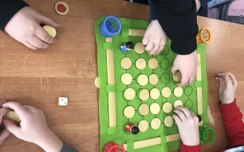 Dzieci grają w grę planszową podczas zajęć korekcyjno kompensacyjnych. Na zielonej macie są drewniane pionki i kostka.