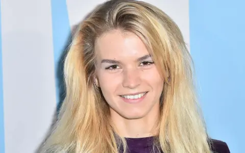 Uśmiechnięta Marta Staśmierska z długimi blond włosami, w fioletowej bluzce, na tle niebiesko-białych pasów.