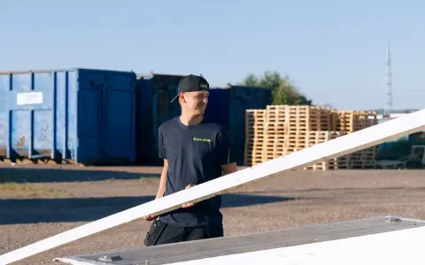 Man i mörkblå t-shirt med "ISOLERAB" logga bär en träskiva. Bakom honom syns blå containrar och träpallar.