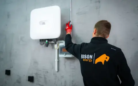 Technik w czerwonych rękawicach mierzy odległość od białego falownika. Działania te mają na celu zapobieganie kradzieży prądu.