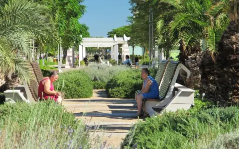 Due donne sedute su panchine in un parco curato, con palme e vegetazione rigogliosa, in una giornata di sole a Riccione centro.