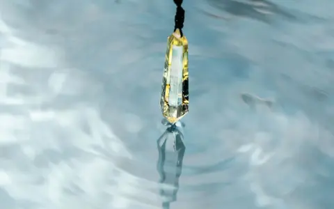 Un cristal de selenita cuelga sobre agua azul, reflejando su brillo. Quizás es porque la selenita necesita agua para purificarse.