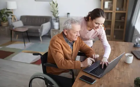 Starszy pan na wózku inwalidzkim z pomocą opiekunki korzysta z laptopa. Nowoczesna technologia wspomagająca ułatwia mu codzienne życie.