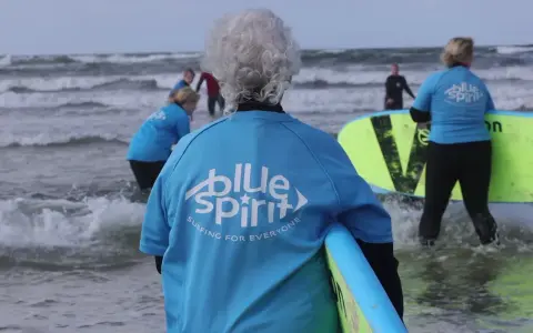 Ältere Dame mit grauem Haar und Surfbrett, bereit für Hvide Sande Surfen. Mehrere Personen in blauen Shirts mit "Blue Spirit" Aufdruck im Wasser.