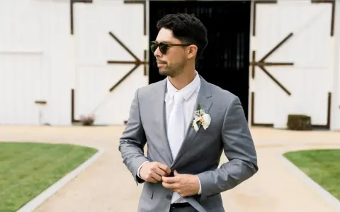Man in a grey suit, white shirt, and tie, looking cool in sunglasses. Perfect for casual men's wedding guest attire in summer.