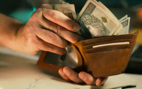 Dłonie wkładają banknoty 100 zł do brązowego portfela. To może być zapłata lub rekompensata.