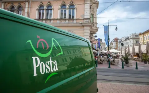 Zielona furgonetka pocztowa z logo "Posta" stoi na ulicy w węgierskim mieście. W tle widać kawiarnie i zabytkowe budynki.
