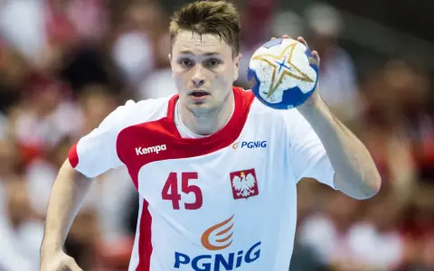 Michał Szyba, numer 45, w białej koszulce z czerwonymi elementami, trzyma piłkę do piłki ręcznej.
