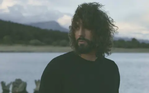 Un hombre con cabello rizado y barba mira hacia el horizonte, entre el miedo y el paraíso de un paisaje acuático y montañoso.