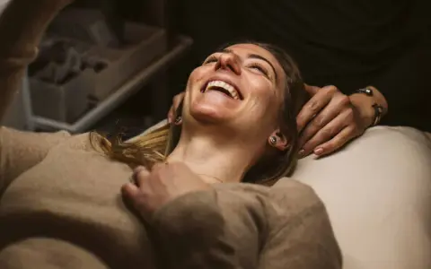 A woman laughs joyfully during a holistic facial treatment, her head tilted back as hands gently support her.