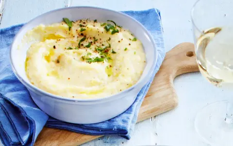 Cremige Polenta, verfeinert mit Butter und Kräutern, serviert in einer Schüssel. Ein Glas Weißwein und ein Holzbrett vervollständigen dieses Polenta Rezept.