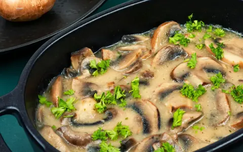 Pyszne pieczarki w śmietanie, doprawione świeżą natką pietruszki, podane w czarnej, owalnej misie.