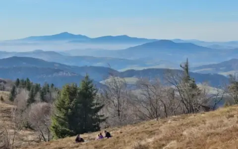 Widok na Beskid Wyspowy z kilkoma osobami odpoczywającymi na zboczu. W oddali widać kolejne pasma górskie w lekkiej mgle.