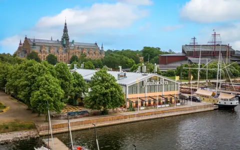 Junibacken, ett museum tillägnat Astrid Lindgren, ligger vid vattnet i Stockholm, med en stor byggnad i bakgrunden och segelfartyg i hamnen.