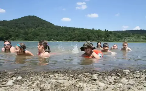Dzieci pluskają się w jeziorze, świetny pomysł na wakacje! W tle zielone wzgórza i błękitne niebo.