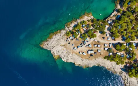 Widok z lotu ptaka na kemping Riviera Makarska. Kamieniste wybrzeże, turkusowa woda i zielone drzewa otaczają liczne przyczepy kempingowe i samochody.