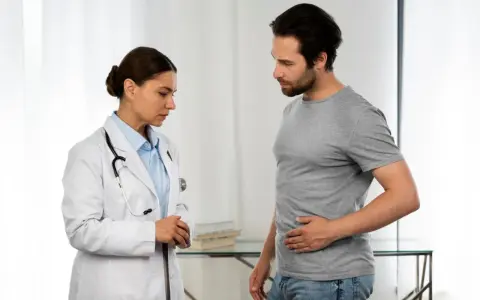 Lekarka w białym kitlu rozmawia z pacjentem, który trzyma się za brzuch. To może być początek rozmowy o tym, jak wygląda badanie urologiczne u mężczyzn.