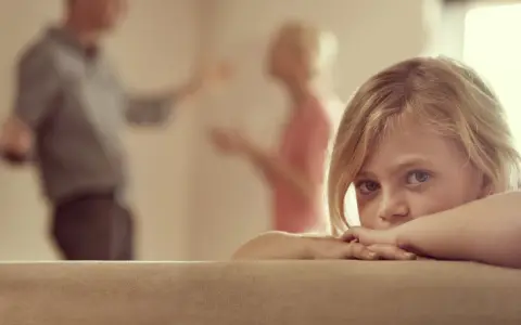 Niña asustada, escondida tras un sofá, mientras sus padres discuten delante de los hijos.