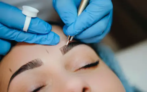 Main d'une esthéticienne en bleu, effectuant un entretien microblading sur les sourcils d'une cliente.