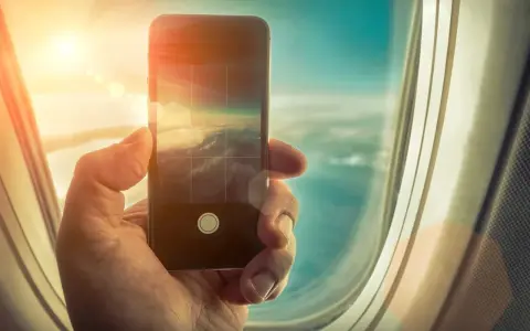 Dłoń trzyma smartfon, fotografując chmury z okna samolotu. Czy w samolocie można używać telefonu? Tak, do robienia zdjęć!