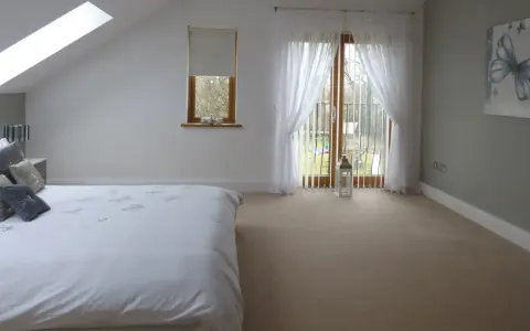 Dormitorio con cama blanca, ventana abuhardillada y puerta acristalada con cortinas. La orientación oeste de los pisos permite una luz suave.