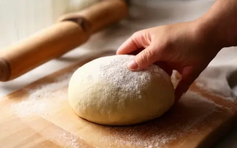 Dłoń dotyka kulki ciasta na pizzę, posypanej mąką. Obok wałek. Czy ciasto na pizzę można mrozić? Tak, można je zamrozić.