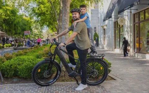 Ojciec z synem na rowerze elektrycznym, który wygląda jak jazda rowerem elektrycznym bez wspomagania. Słońce, zieleń i sklepy w tle.