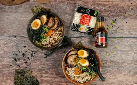 Dwa miski aromatycznego ramenu z boczkiem, jajkiem i nori. Obok opakowanie makaronu i sosu.