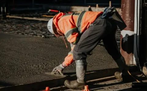 Obrero con casco y chaleco naranja trabajando con hormigón autocompactante, alisando la superficie con una llana.