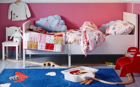 Habitación infantil con dos camas blancas, cojines de nube y mantas coloridas. Alfombra azul con tren de juguete y vías.