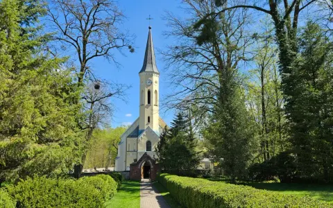 Jasny kościół z wieżą zegarową otoczony zielenią. W oddali widać drzewa, a ścieżka prowadzi do wejścia. To miejsce, gdzie czas płynie spokojnie, jak w dzień świętopietrze.