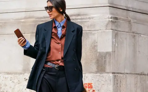 Mujer con estilo combinando traje negro con camisa marrón y azul, gafas de sol y móvil en mano.