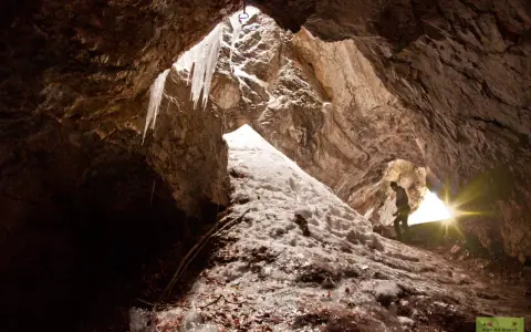Wnętrze jaskini z lodowymi sopelkami i człowiekiem wspinającym się po oblodzonym zboczu. To prawdziwa dolina ku dziurze, gdzie światło przebija się przez otwór.