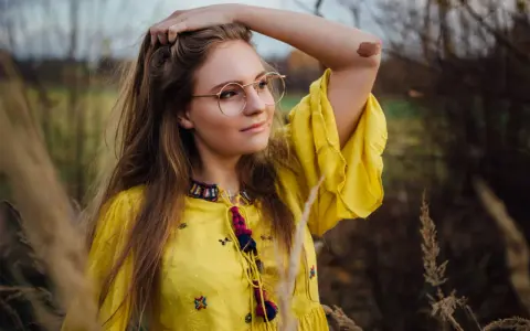 Portret dziewczyny w okularach i żółtej sukience, wśród traw. Różne rodzaje portretów w fotografii mogą uchwycić taką naturalną, swobodną chwilę.