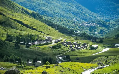 Un villaggio alpino nel **Pian della Regina**, con case in pietra e un fiume che scorre tra prati verdi e boschi lussureggianti.