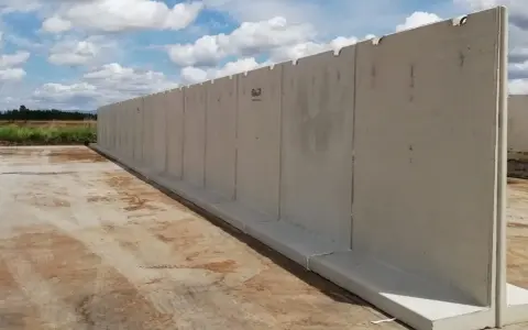Larga pared de separador de hormigón prefabricado, instalada sobre una base de hormigón, con un cielo azul y nubes blancas al fondo.
