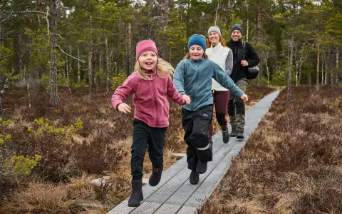 Familj njuter av aktiviteter i skogen med barn på en spång. Två barn springer glatt framför sina föräldrar i en vacker skogsmiljö.