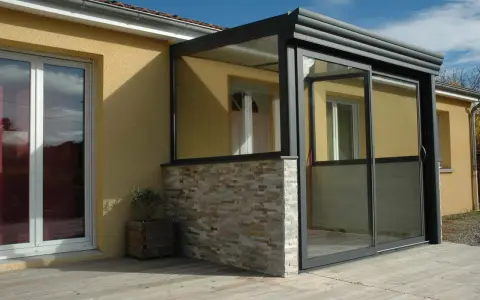 Une véranda moderne avec murs en pierre et grandes baies vitrées permet une entrée directe dans le séjour.