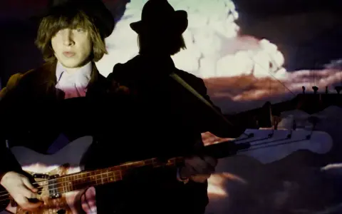 Jacco Gardner, con sombrero y chaqueta, toca el bajo frente a una proyección psicodélica.