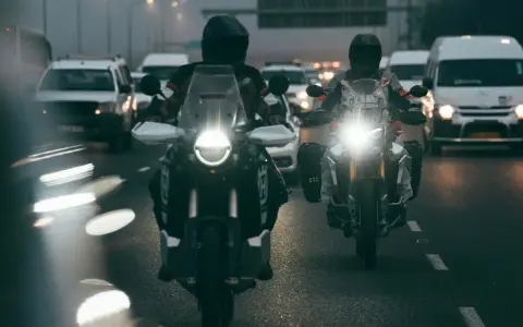 Dwóch motocyklistów w kaskach jedzie w deszczu. Ich motocykle mają włączone światła, a droga polna jest mokra i błyszcząca.