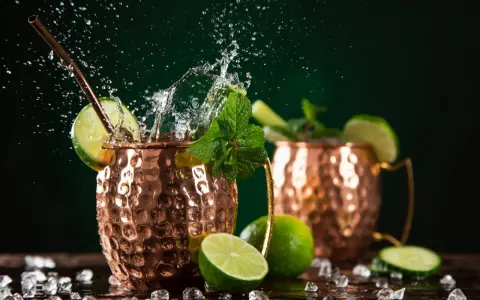 Dwa miedziane kubki z orzeźwiającym Moscow Mule, ozdobione limonką i miętą, z bryzgami wody i kostkami lodu.
