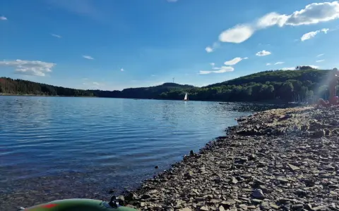 Sonniger Tag am See in NRW. Ein Segelboot gleitet über das Wasser, während am steinigen Ufer Menschen entspannen.