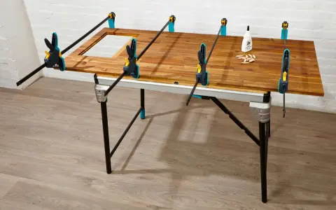 Holzplatte wird mit Zwingen fixiert, um einen Tisch selber bauen. Kleber und Dübel liegen bereit.
