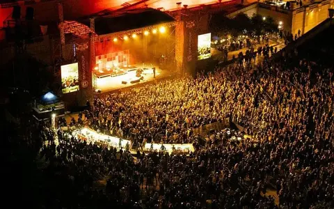 Multitudinaria asistencia a conciertos en julio. Una banda toca en un escenario iluminado ante miles de fans.