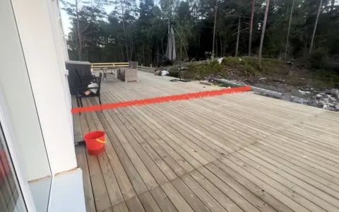 Nybyggd altan med en röd markering som visar var ett vindskydd ska byggas. Trädgårdsmöbler och en parasoll finns på plats.