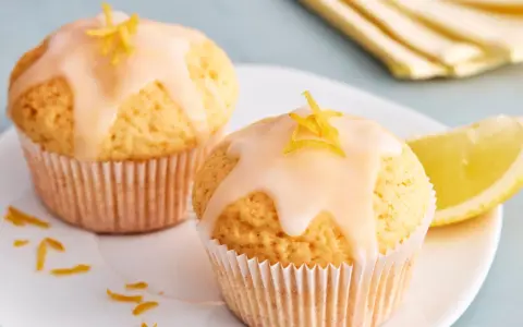 Zwei einfache Zitronenmuffins mit Zuckerguss und Zitronenschale garniert, daneben ein Zitronenschnitz.