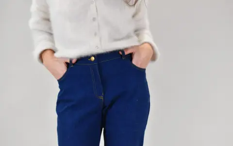 Gros plan sur une personne portant un pull blanc et un pantalon en jean bleu avec les mains dans les poches.