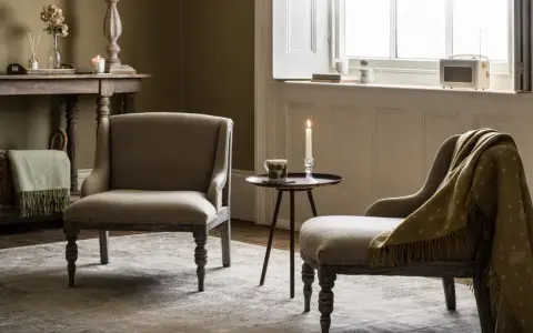 Przytulny salon w stylu angielskim. Dwa fotele, stolik z zapaloną świecą, kosz z pledem i stylowy mebel pod oknem.