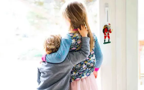 Zwei Kinder umarmen sich vor einem Fenster. Das jüngere Kind, das Angst hat, wird vom älteren Kind getröstet. Ein Weihnachtsmann-Anhänger hängt an der Tür.